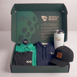 An open branded box containing a green patterned shirt, a navy hoodie, a black graphic T-shirt, a white insulated tumbler, and a black baseball cap with a brown patch, all arranged on a light gray background.