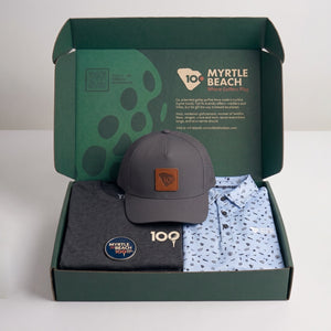 A green branded box opened with a grey graphic t-shirt, a grey cap and a blue printed polo shirt.