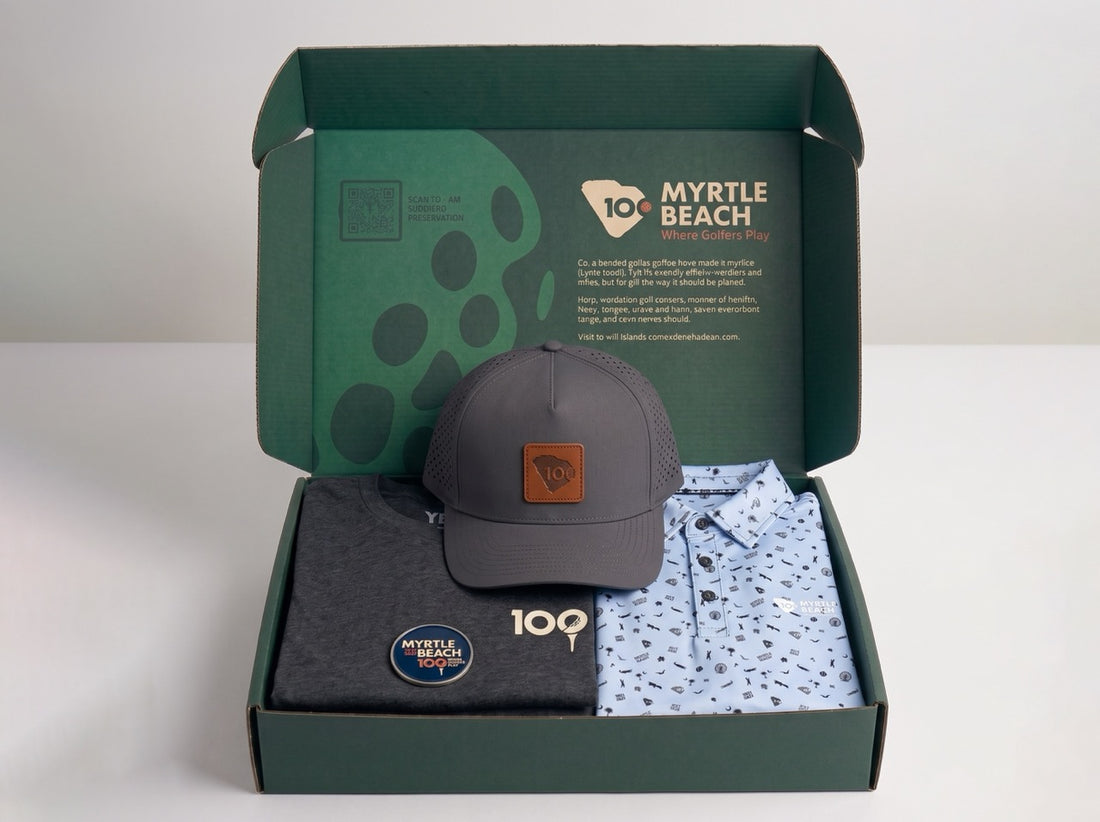A curated gift set in an open dark green box from 100 Myrtle Beach, featuring a dark gray baseball cap with a brown leather patch, a dark gray t-shirt, and a light blue patterned button-down shirt.