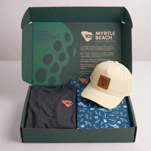 Open dark green gift box containing a dark gray t-shirt, a blue patterned polo, and a cream baseball cap with a brown patch. The inside lid shows 'MYRTLE BEACH' branding and text.
