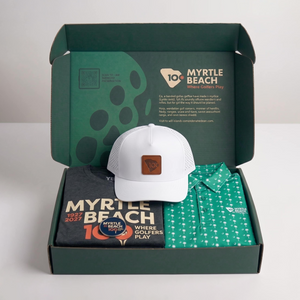 A curated gift set in an open dark green box, featuring a white trucker hat with a brown leather patch, a dark gray t-shirt, and a green patterned polo shirt.